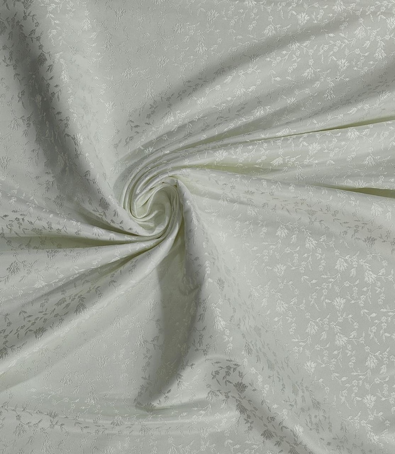 May include: Ivory-colored fabric with a subtle floral pattern. The fabric is draped, creating soft folds and shadows. The delicate design adds texture and visual interest to the material, suitable for apparel or home decor projects.
