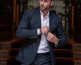 May include: A man wearing a dark blue suit jacket and matching pants. He is wearing a white dress shirt with a button-down collar and a dark blue pocket square. He is also wearing a watch on his left wrist and a bracelet on his right wrist.