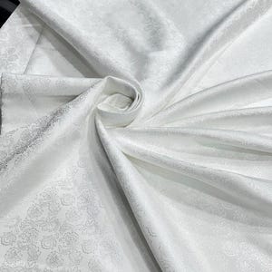May include: White fabric with a floral pattern. The fabric is folded and draped, creating soft shadows and highlights. The floral design is subtle, adding texture to the smooth, satiny material. The fabric appears to be a luxurious textile, suitable for various applications.