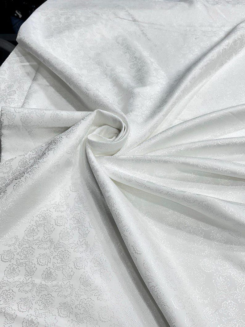May include: White fabric with a floral pattern. The fabric is folded and draped, creating soft shadows and highlights. The floral design is subtle, adding texture to the smooth, satiny material. The fabric appears to be a luxurious textile, suitable for various applications.