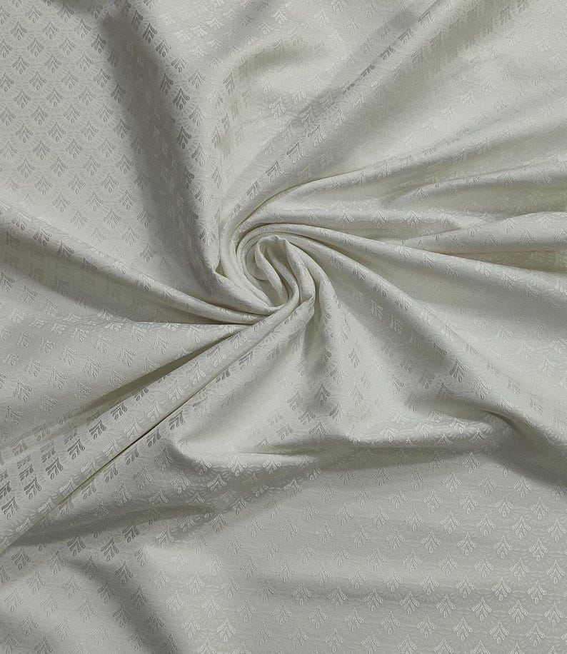 May include: Off-white fabric with a subtle, repeating geometric pattern. The fabric is draped and folded, creating soft shadows and highlights. The texture appears smooth with a slight sheen, suggesting a luxurious material.
