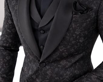 May include: A black tuxedo jacket and vest set featuring a tonal floral jacquard pattern, a solid black satin shawl lapel, a matching black shirt, a patterned bow tie, a pocket square, and a gold-tone wristwatch with a black dial.