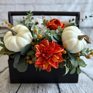 May include: A black wooden box with a handle filled with a fall floral arrangement. The arrangement includes white pumpkins, orange and red dahlias, and green foliage.