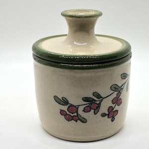 May include: A small, white ceramic jar with a green lid and a painted design of red berries and green leaves. The jar has a simple, rustic style.