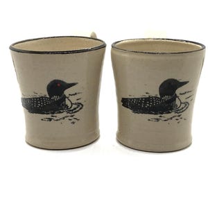 May include: Two ceramic mugs with a black and white loon design. The mugs are a light beige color with a dark brown rim.