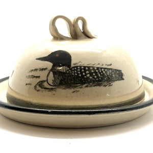 May include: A ceramic butter dish with a black and white loon design. The dish has a lid with two handles and a black rim.
