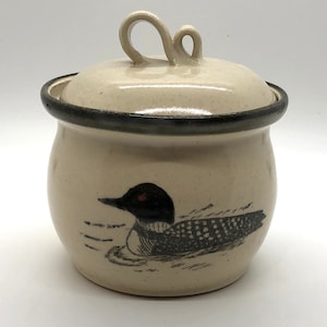 May include: A white ceramic jar with a black rim and a lid with a loop handle. The jar has a black and white illustration of a loon on the front.