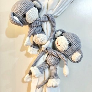 May include: Two gray and white crocheted elephant curtain tiebacks. The elephants have large ears, white tusks, and red smiles. The tiebacks are wrapped around a white curtain.
