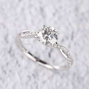 Puede incluir: Un anillo de compromiso de plata con una piedra central redonda y clara y una banda retorcida adornada con piedras más pequeñas.