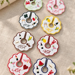 May include: Eight circular white fabric wine glass markers with scalloped edges and embroidered motifs. Each marker features a central hole, a slit, and unique colourful designs like fruits, drinks, and initials. The borders are stitched in various colours.