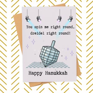 May include: Hanukkah greeting card with a dreidel illustration. The card features the text "You spin me right round, dreidel right round!" and "Happy Hanukkah". The card is on a kraft paper envelope with a gold chevron pattern.