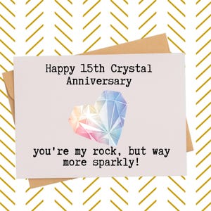 May include: A white greeting card with a brown envelope featuring the text Happy 15th Crystal Anniversary and you're my rock, but way more sparkly! in black font. A colorful, faceted heart-shaped crystal graphic is centered on the card.