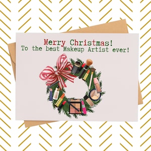 May include: A Christmas greeting card with a makeup theme. The card features a wreath made of makeup brushes, lipsticks, and other beauty products. The text reads "Merry Christmas! To the best Makeup Artist ever!" The card is on a white background with a gold chevron pattern.