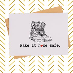 May include: A white greeting card with a drawing of combat boots. The text "Make it ❤️ home safe" is printed below the boots. The card is inside a brown envelope, with a gold and white chevron pattern in the background.