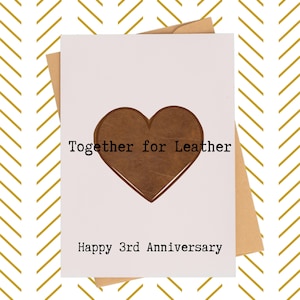 May include: A white greeting card with a brown leather heart cutout and the text "Together for Leather" and "Happy 3rd Anniversary". The card is inside a brown envelope, set against a gold and white chevron pattern.