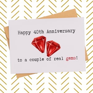 May include: A white greeting card with the text "Happy 40th Anniversary" and "to a couple of real gems!" features two red gem illustrations. The card is on a brown envelope, set against a gold and white chevron pattern.