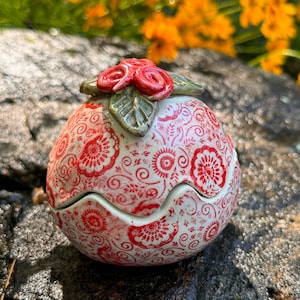 May include: A white ceramic trinket box with a red and white floral pattern. The lid is shaped like a flower with red petals and green leaves.
