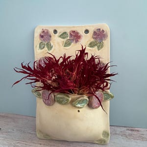 May include: A cream-colored ceramic wall pocket planter featuring a rectangular backplate with two holes for hanging. The surface is decorated with sculpted purple flowers and green leaves, and it contains a cluster of dark red dried foliage.