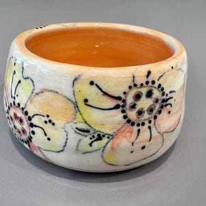 May include: A ceramic bowl with a floral design. The bowl is white with yellow, pink, and black floral patterns. The interior of the bowl is orange. The bowl is round and has a thick rim.