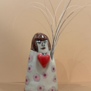 May include: A ceramic vase in the shape of a figure with brown hair and a red heart. The vase is white with pink and purple polka dots. Tall, thin, light-colored stems extend from the top of the vase.