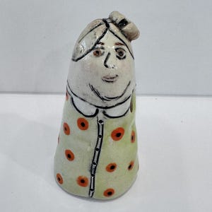 May include: A whimsical ceramic figurine of a woman with a smiling face, wearing a light green dress with orange polka dots. The figurine has a white collar and a small bow on top of her head. The figure is hand-painted.