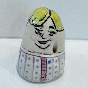 May include: A hand-painted ceramic thimble featuring a whimsical face with yellow hair and a smiling expression. The thimble's base has a striped pattern with blue polka dots and a vertical purple stripe design.