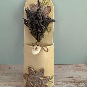 May include: A cream-colored ceramic wall pocket featuring a sculpted flower design at the base and top. A bundle of dried purple lavender is tucked inside, tied with twine and a small white oval tag displaying a brown illustration of a mouse.