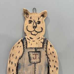 May include: A flat, wall-hanging ceramic plaque shaped like a cat wearing blue-tinted overalls. The cat has a cream-colored surface with black etched outlines and markings. A pocket on the overalls features a red letter J. A black wire loop is at the top.