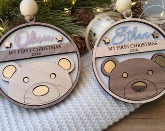 May include: Two circular wooden ornaments featuring a bear face design. One ornament is engraved with the name Olivia and the other with Ethan. Both include the text MY FIRST CHRISTMAS 2026, small star accents, a wooden bead, and a beige ribbon hanger.