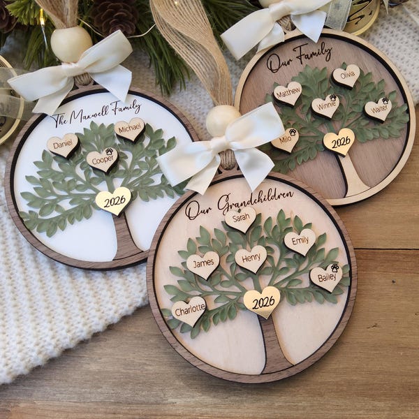 Puede incluir: Tres adornos circulares de madera con un diseño de árbol cortado a láser con hojas verdes. Cada adorno muestra pequeños corazones de madera grabados con nombres y el año 2026, rematados con un lazo de cinta blanca y una cuenta de madera.