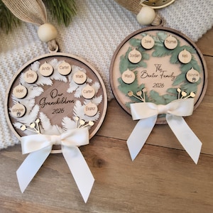 May include: Two circular wooden ornaments featuring a wreath design, white ribbon bows, and hanging loops with wooden beads. One ornament displays the text Our Grandchildren 2026 with names on small wooden circles. The other shows The Baxter Family 2026.