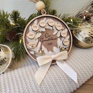 May include: A circular wooden ornament featuring a white wreath design, the text Our Grandchildren 2026, and small wooden discs engraved with names. The ornament is accented with a cream-coloured ribbon bow and a wooden bead at the top hanging loop.