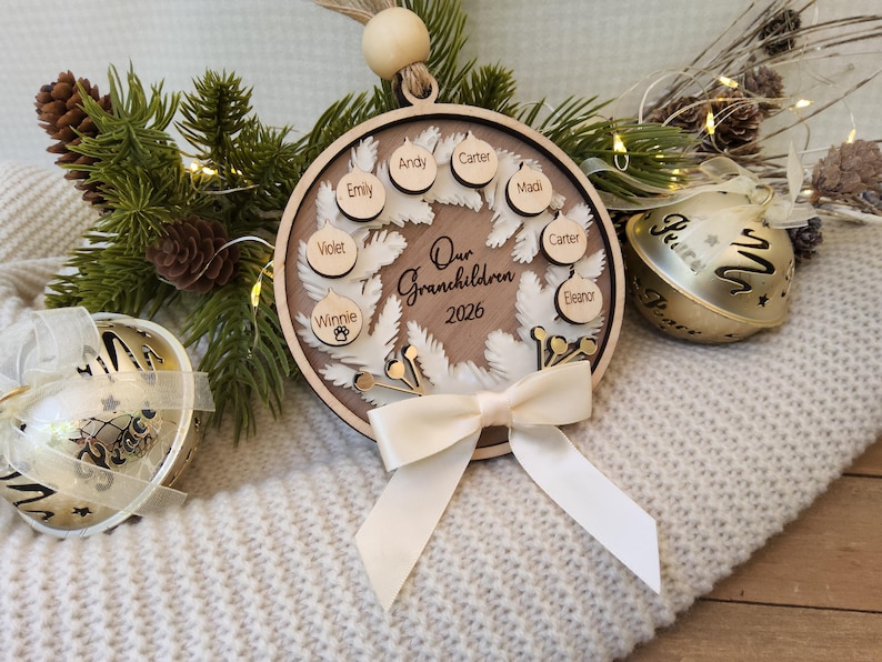 May include: A circular wooden ornament featuring a white wreath design, the text Our Grandchildren 2026, and small wooden discs engraved with names. The ornament is accented with a cream-coloured ribbon bow and a wooden bead at the top hanging loop.