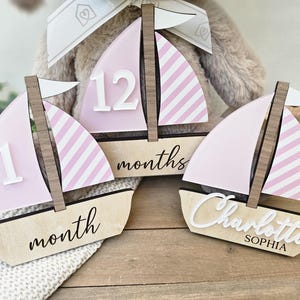 May include: Wooden sailboat-shaped milestone markers. The sails are pink with white stripes, and the hulls are natural wood. One marker has the number "1" and the word "month", another has "12" and "months", and the third has the name "Charlotte Sophia".