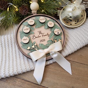 May include: A round wooden Christmas ornament featuring a green wreath design, a cream-coloured centre with the text The Baxter Family 2026, and six small circular wooden tags with names attached. A cream satin ribbon bow is tied at the bottom.
