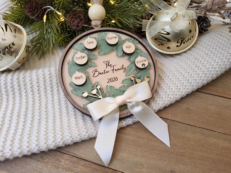 May include: A round wooden Christmas ornament featuring a green wreath design, a cream-coloured centre with the text The Baxter Family 2026, and six small circular wooden tags with names attached. A cream satin ribbon bow is tied at the bottom.