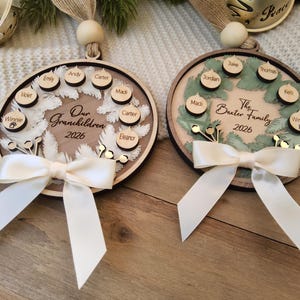 May include: Two round wooden ornaments featuring a wreath design, a cream-coloured satin bow, and a hanging loop with a wooden bead. The left ornament reads Our Grandchildren 2026 with names on small wooden discs. The right reads The Baxter Family 2026.