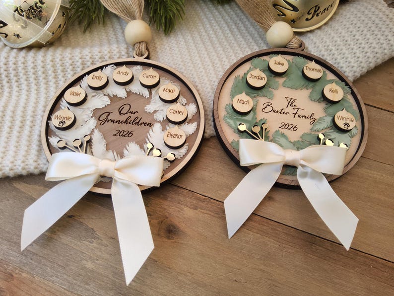 May include: Two round wooden ornaments featuring a wreath design, a cream-coloured satin bow, and a hanging loop with a wooden bead. The left ornament reads Our Grandchildren 2026 with names on small wooden discs. The right reads The Baxter Family 2026.