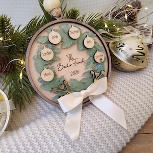 May include: A round wooden Christmas ornament featuring a layered green wreath design. The centre is engraved with The Baxter Family 2026. Six small wooden bauble cutouts are attached, each engraved with a name: Jake, Thomas, Kelli, Winnie, Madi, and Jordan. A white ribbon bow is tied at the bottom.