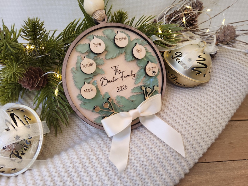 May include: A round wooden Christmas ornament featuring a layered green wreath design. The centre is engraved with The Baxter Family 2026. Six small wooden bauble cutouts are attached, each engraved with a name: Jake, Thomas, Kelli, Winnie, Madi, and Jordan. A white ribbon bow is tied at the bottom.