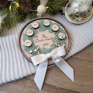 May include: A round wooden Christmas ornament featuring a green wreath design with small circular wooden tags engraved with the names Thomas, Kelli, Winnie, Jordan, Jake, and Madi. The centre text reads The Baxter Family 2026, accented by a white ribbon.