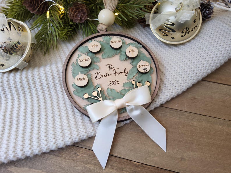 May include: A round wooden Christmas ornament featuring a green wreath design with small circular wooden tags engraved with the names Thomas, Kelli, Winnie, Jordan, Jake, and Madi. The centre text reads The Baxter Family 2026, accented by a white ribbon.