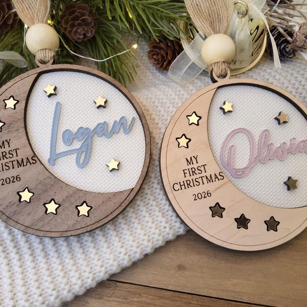 Puede incluir: Dos adornos circulares de madera con diseño de media luna. Uno es de madera oscura con el nombre Logan en azul, el otro de madera clara con el nombre Olivia en rosa. Ambos incluyen el texto My First Christmas 2026 y pequeñas estrellas.