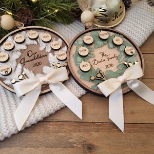 May include: Two circular wooden ornaments featuring a wreath design. One reads Our Grandchildren 2026, the other The Baxter Family 2026. Both include small wooden name tags, a cream-coloured ribbon bow, and a wooden bead at the top for hanging.