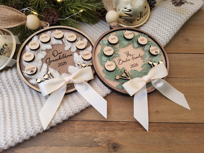 May include: Two circular wooden ornaments featuring a wreath design. One reads Our Grandchildren 2026, the other The Baxter Family 2026. Both include small wooden name tags, a cream-coloured ribbon bow, and a wooden bead at the top for hanging.