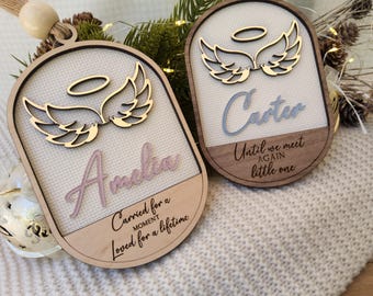 May include: Two oval wooden ornaments featuring a layered design with a cream-colored textured background. Each ornament displays a gold-toned halo and angel wings above a name, Amelia or Carter, and the phrases Carried for a moment, Loved for a lifetime or Until we meet again little one.