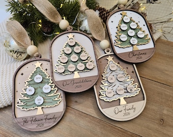 May include: Four oval wooden Christmas ornaments featuring layered tree designs with circular name tags. Each ornament includes a tan ribbon, a wooden bead, and engraved text such as The Filmore Family 2026, Our Grandchildren 2026, Our Family 2026, and Christmas 2026.