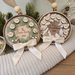 May include: Two circular wooden ornaments featuring layered wreath designs, cream ribbons, and hanging beads. One displays The Baxter Family 2026 with names on small wooden tags, while the other reads Our Grandchildren 2026 with names on similar tags.