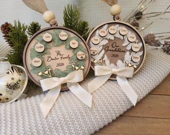 May include: Two circular wooden ornaments featuring layered wreath designs, cream ribbons, and hanging beads. One displays The Baxter Family 2026 with names on small wooden tags, while the other reads Our Grandchildren 2026 with names on similar tags.