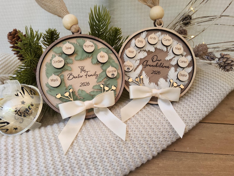 May include: Two circular wooden ornaments featuring layered wreath designs, cream ribbons, and hanging beads. One displays The Baxter Family 2026 with names on small wooden tags, while the other reads Our Grandchildren 2026 with names on similar tags.